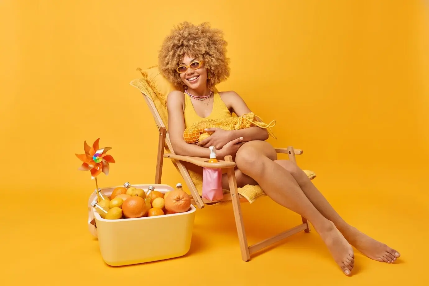 Vrolijke vrouw houdt een netzak met citrusvruchten vast, kijkt blij weg en zit op een ligstoel, gekleed in een badpak en een zonnebril; geniet van een fijne zomerdag en een perfecte vakantie, geïsoleerd tegen een gele muur, met een draagbare koelbox in de buurt.