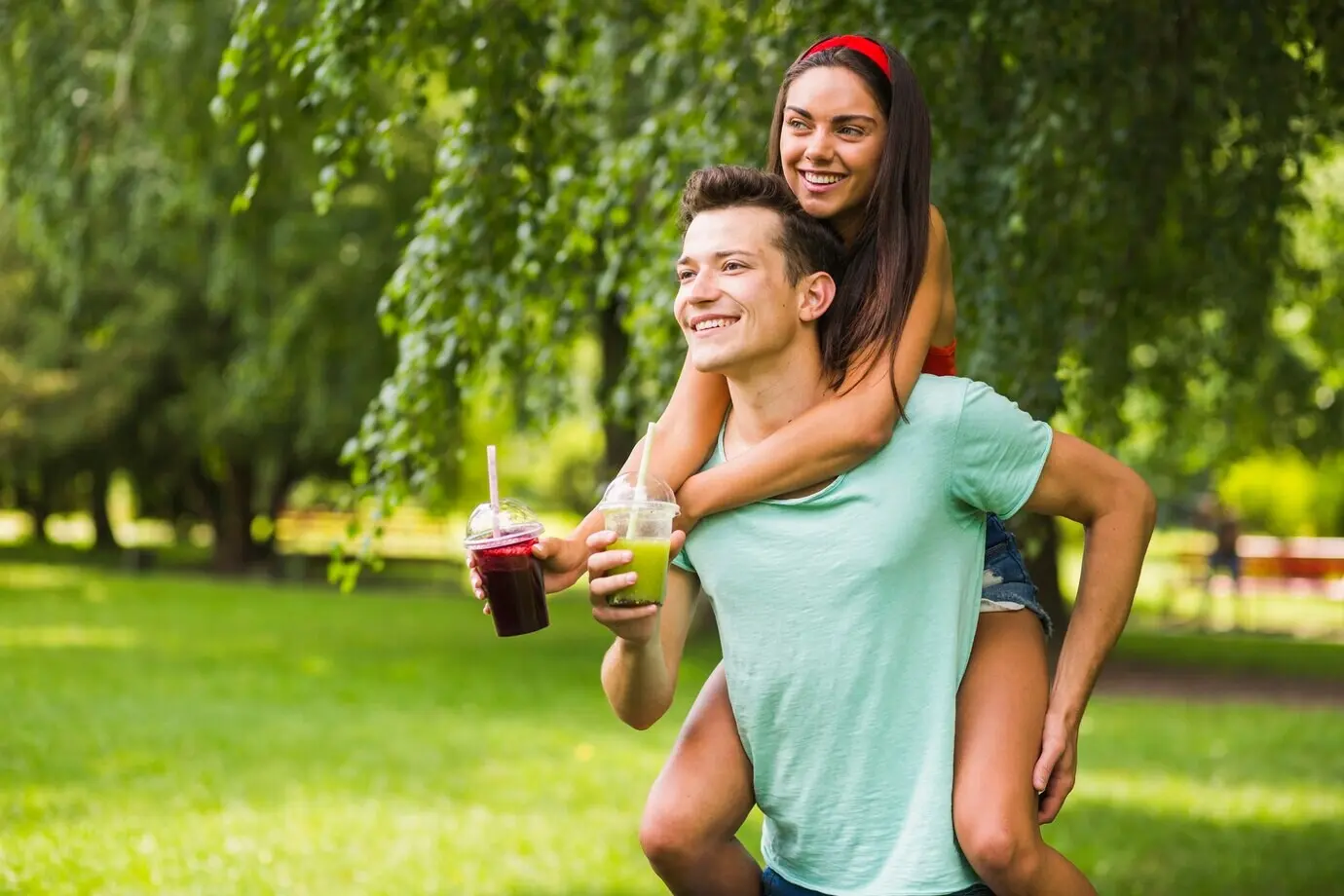 Een man draagt zijn vriendin op zijn rug en houdt smoothies vast in het park.