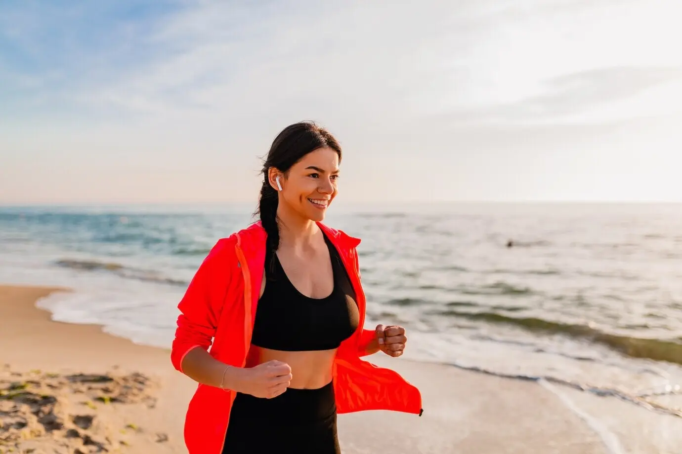 Jonge, aantrekkelijke, slanke vrouw die ’s ochtends bij zonsopgang sportoefeningen doet en jogt op het strand aan zee, in sportkleding, met een gezonde levensstijl, luisterend naar muziek via oordopjes, gekleed in een roze windjack.